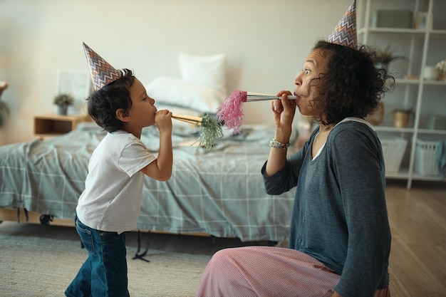Side view shot of cheerful young mixed race female and her cute little son wearing conical hats, blowing whistle while celebrating birthday at home alone during quarantine