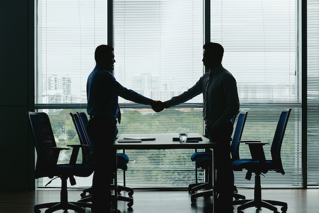 Side view of silhouettes of two unrecognizable men shaking hands in the office