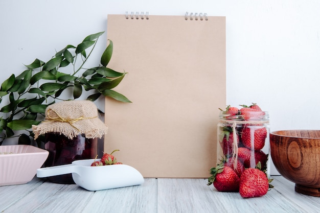 Side view of a sketchbook with strawberry jam in a glass jar and fresh ripe strawberries in a glass jar on rustic