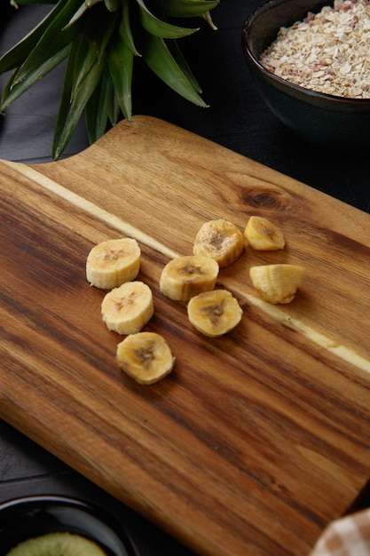 Free photo side view of sliced banana on cutting board with pineapple leaves and oats on black background