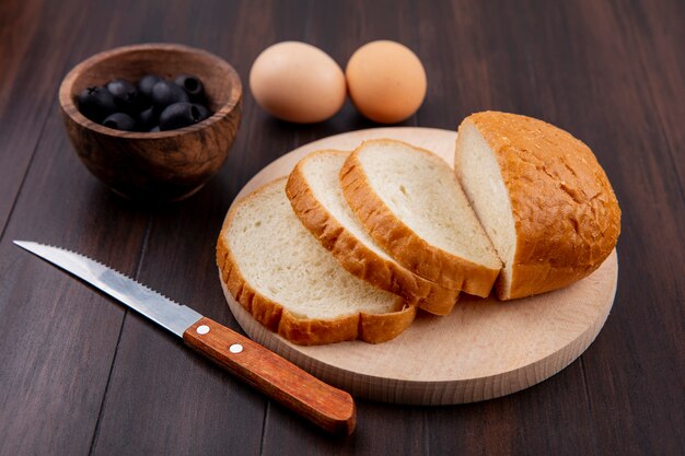 Side view of sliced bread on cutting board and knife with eggs and bowl of black olive on wood