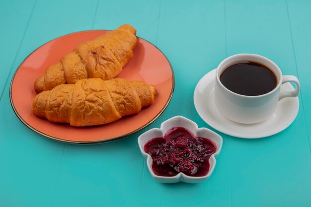 Side view of sliced croissants and raspberry jam with cup of tea on blue background