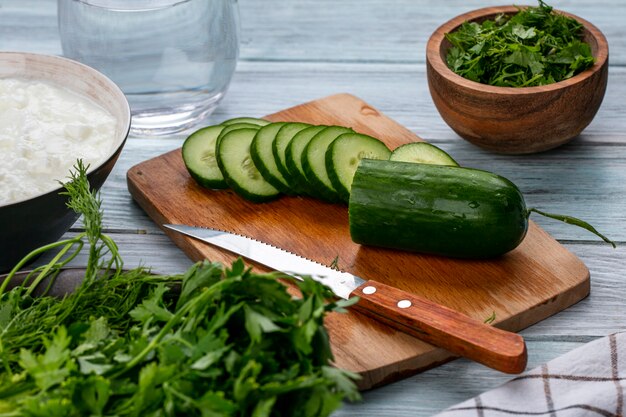 Side view of sliced cucumbers on a board with a knife and greens on a gray surface