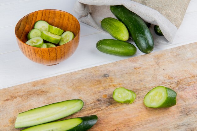 Side view of sliced and cut cucumber on cutting board with bowl of cucumber slices and cucumbers spilling out of sack on wooden table