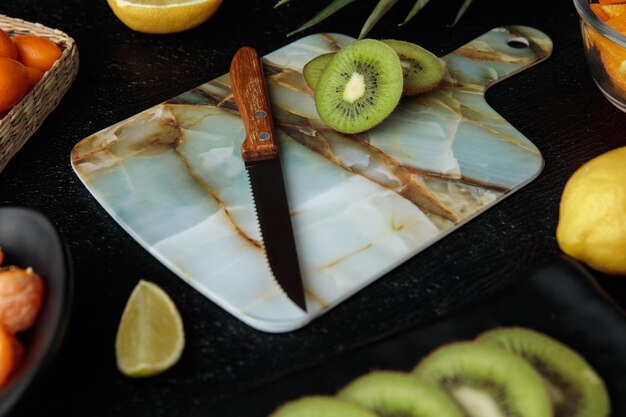 Side view of sliced kiwi and knife on cutting board with other fruits around on black background