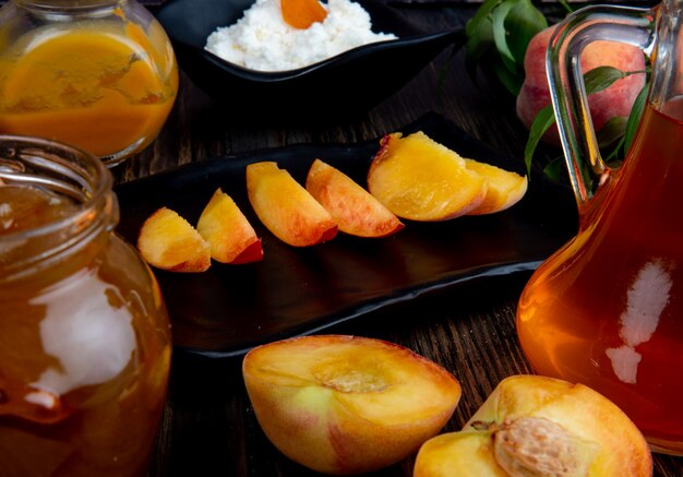 Side view of sliced peach on a black tray and a glass jar with peach jam on rustic wooden background