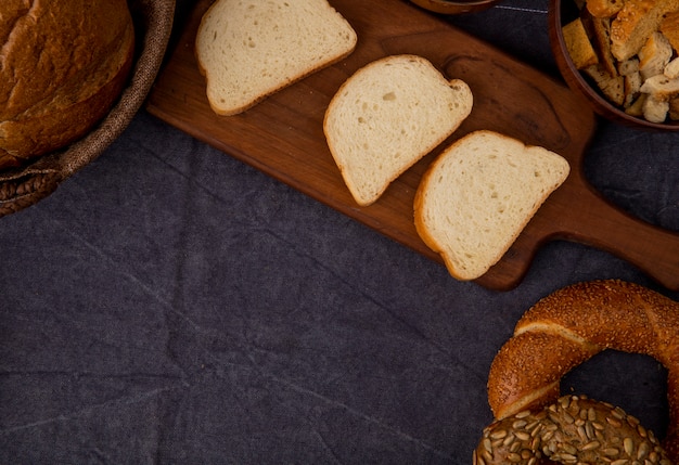Side view of sliced white bread on cutting board with bagels and bread pieces on maroon background with copy space