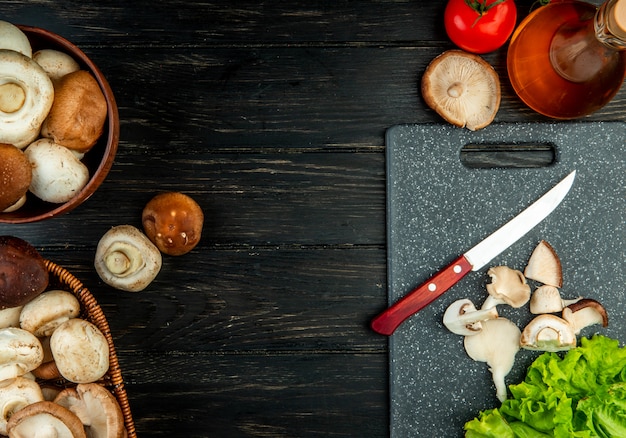 Side view of sliced and whole mushrooms with kitchen knife on a black cutting board on black wood with copy space