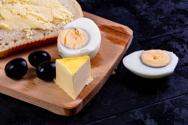 Side view slices of bread and butter on board with olives and boiled egg on black