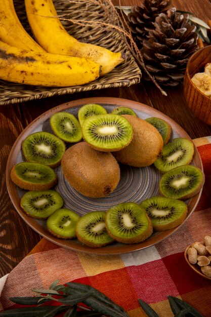 Вид сбоку ломтиков киви на тарелку и гроздь бананов в плетеной корзине на дереве
