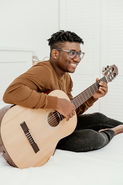 Улыбающийся музыкант мужского пола, играющий на гитаре на кровати, вид сбоку