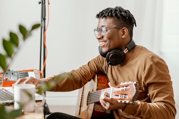 Side view of smiley male musician at home playing guitar and mixing with laptop