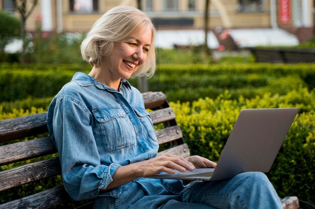 Side view of smiley older woman outdoors on bench with laptop