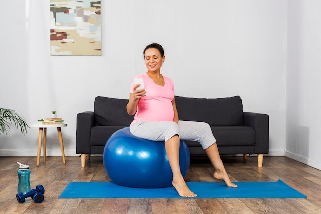 Side view of smiley pregnant woman at home using smartphone and training with ball