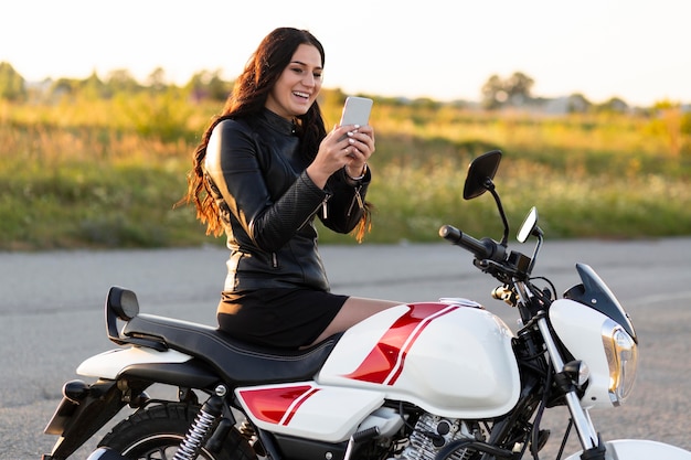 Foto gratuita vista laterale della donna sorridente guardando smartphone mentre è seduto sulla sua moto