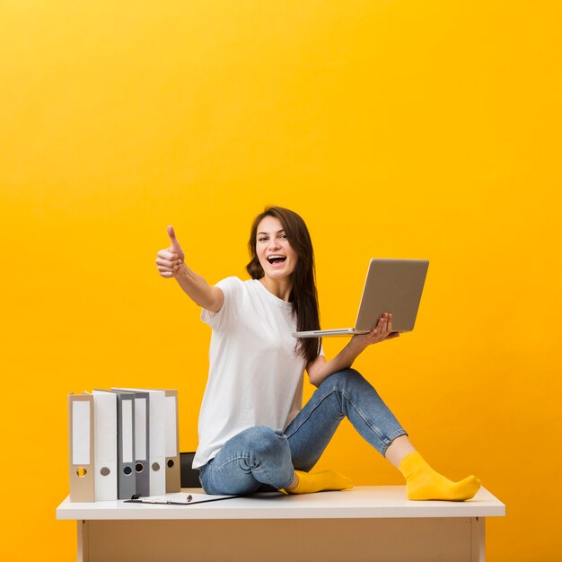 Side view of smiley woman sitting on desk and giving thumbs up while holding laptop