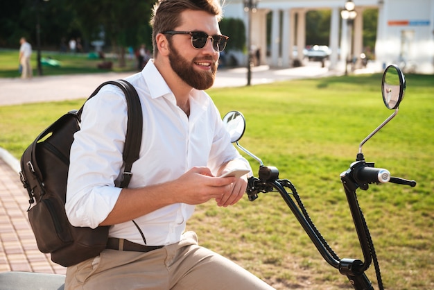 Side view of smiling bearded man in sunglasses sitting on modern motorbike outdoors with smartphone and looking away