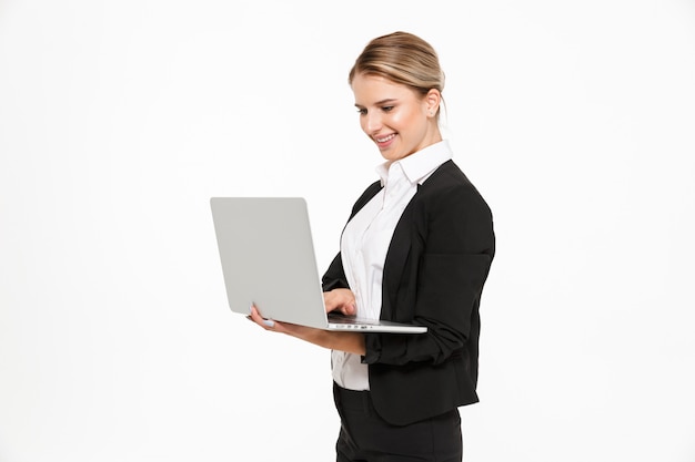 Side view of smiling blonde business woman holding and using laptop computer over white wall