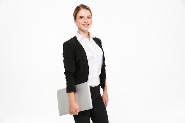 Side view of smiling blonde business woman posing in studio with laptop computer in hand and  over white wall