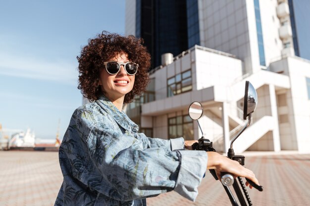 Side view of smiling curly woman in sunglasses posing on modern motorbike outdoors and looking away