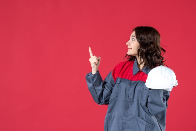 Side view of smiling female architect holding hard hat and pointing up on isolated red background