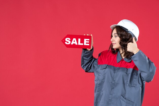 ハード帽子をかぶって、孤立した赤い背景でOKジェスチャーを作る販売アイコンを指している制服を着て笑顔の女性労働者の側面図