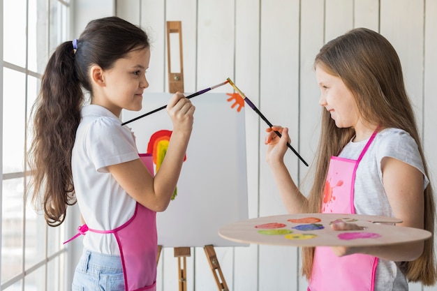 Side view of smiling two girls touching their paint brushes while painting on canvas