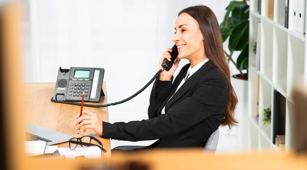 Side view of a smiling young woman sitting in office talking on telephone