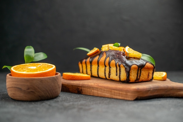 Side view of soft cakes on cutting board and cut orangeswith leaves on dark table