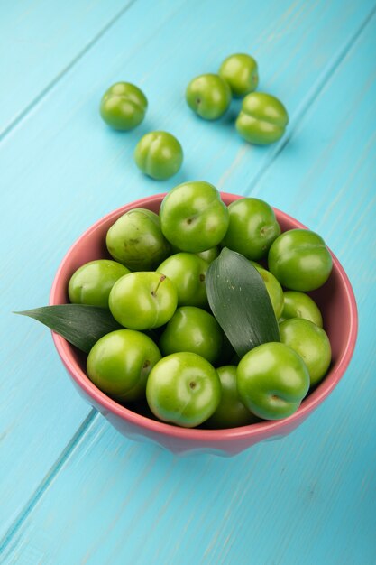 Side view of sour green plums in a bowl on blue wooden table