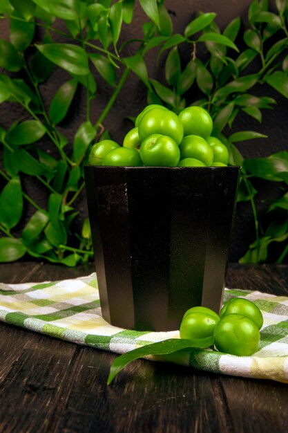 Side view of sour green plums in a bowl on plaid napkin on wooden surface at green leaves table