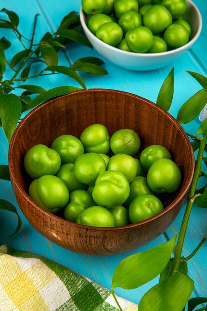 Side view of sour green plums in bowls on blue wooden table