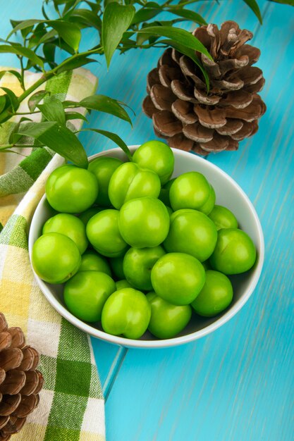 Side view of sour green plums in a white bowl and cones on plaid fabric on blue table table