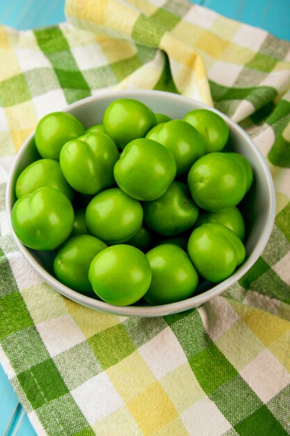 Side view of sour green plums in a white bowl on plaid fabric table