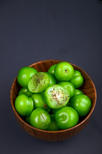 Side view of sour green plums in a wooden bowl on black table