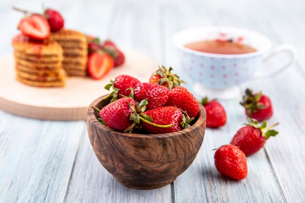 Side view of strawberries in bowl with tea and waffle biscuits on wood