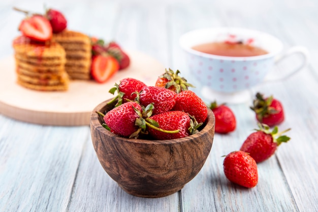 Side view of strawberries in bowl with tea and waffle biscuits on wood