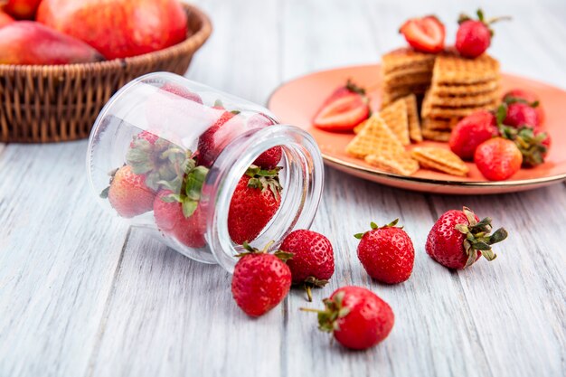 Side view of strawberries spilling out of jar and strawberry cake in plate with fruits on wood
