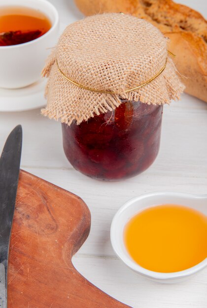 side view of strawberry jam in jar with melted butter cup of tea knife on cutting board and baguette on wooden table