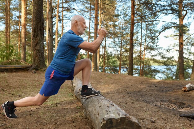 Вид сбоку сильного подтянутого старшего мужчины с бородой, тренирующегося в лесу, выпады doig, держа ноги на бревне. Сосредоточенный пожилой мужчина делает физические упражнения для мышц ног в солнечный летний день