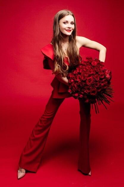 세련된 빨간 드레스를 입은 긴 머리를 한 멋진 젊은 갈색 머리의 측면 스튜디오 초상화 그녀는 손에 든 큰 빨간 장미를 보고 웃고 있습니다.