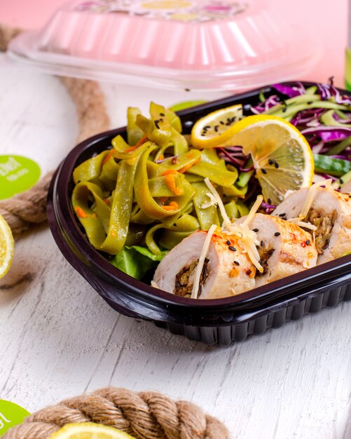 Side view of stuffed chicken roll with vegetable garlic and nuts served with cabbage salad and lemon slice in delivery box