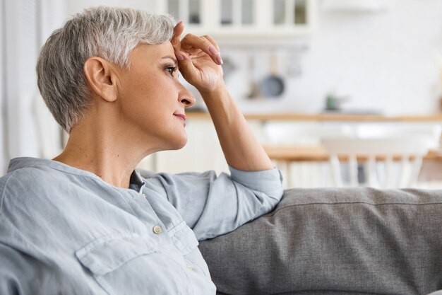 Side view of stylish elderly mature woman with neat make up and short hair relaxing on sofa at home, having thoughtful dreamy facial expression. Retired female sitting on couch in living room