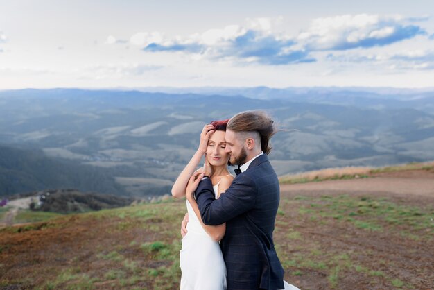 Side view of stylish guy hugs a red-haired girl in a field and the wind develops their hair