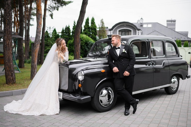 Вид сбоку на стильного и красивого жениха в черном смокинге, опирающегося на ретро-автомобиль и смотрящего на невесту, которая стоит напротив и мило смотрит на него во время свадебной прогулки на открытом воздухе