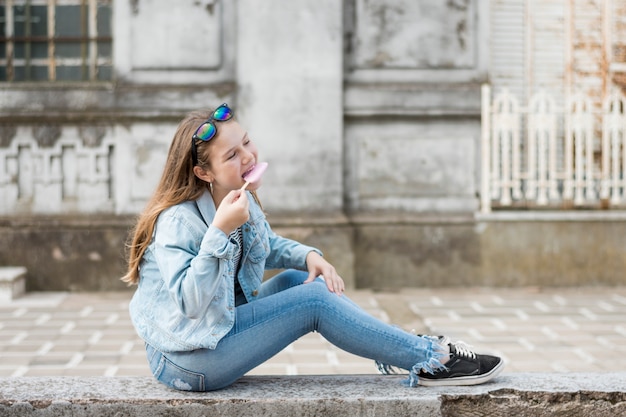 Foto gratuita vista laterale dell'adolescente alla moda che ubicazione sulla parete che mangia gelato