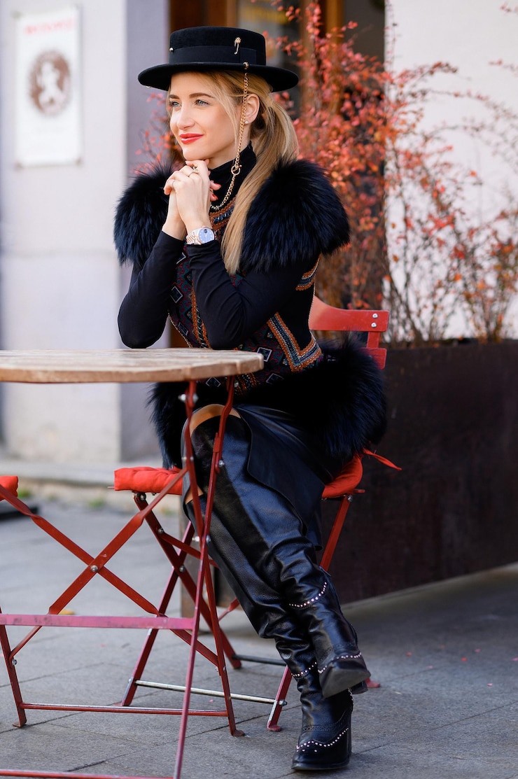 Free Photo side view of stylish young girl with tied hair and black fedora on head dressed in vogue outfit and leather footwear keeping hands together and looking away while resting in outdoor cafe