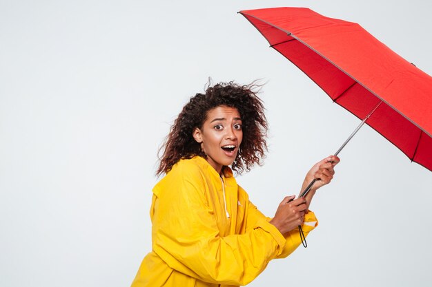 Side view of surpised african woman in raincoat holding umbrella