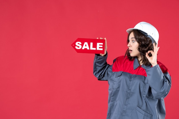 ハード帽子をかぶって、孤立した赤い背景に眼鏡のジェスチャーを作る販売アイコンを指している制服を着た驚いた女性労働者の側面図