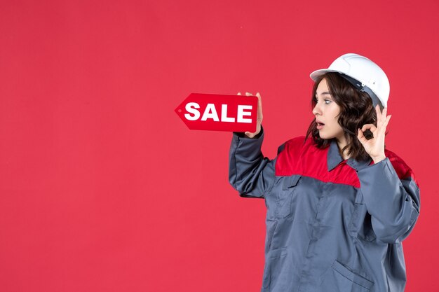 Side view of surprised female worker in uniform wearing hard hat and pointing sale icon making eyeglasses gesture on isolated red background